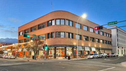 A modern brick building with large windows, featuring a food hall, at a street intersection during twilight.